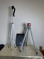 Two silver portable tripods shown side by side against a wall, one with a black handle and one with a flat red mount top plate, on a covered surface.