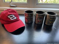 Red Dale Jr #8 baseball cap seen next to three cylindrical metal beer coolers featuring NASCAR and Indianapolis Motor Speedway logos.