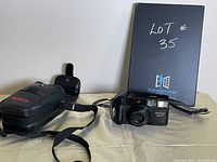 Full view of Pentax Zoom 90 camera with accompanying carrying case and adjustable wrist strap, displayed on a light surface with a black notebook in the background.