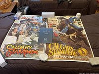 Overview of lot including two Calgary Stampede posters from 1976 and one from 1977 on a black surface, showing colorful rodeo scenes and event dates.