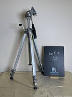 Velbon VG-3 tripod standing on floor against a wall with a black sign indicating lot number 32, showing full view with tripod legs retracted and extended, head and adjustment handle visible.