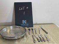 Vintage silver plate round tray with nine vintage silver plated butter spreaders laid out on a table with a black box behind them.