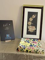 Photo shows the framed pressed flower piece standing on a yellow floral surface with a black lot number sign next to it.