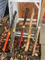 Three sledgehammers and two axes resting against a 6-foot aluminum step ladder outside on leaves and gravel.