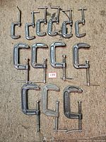 Top view of 15 metal C-clamps arranged on carpet surface. Sizes vary from small to large, cleaned condition with metallic finish.