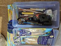 Multiple assorted paint brushes, some in plastic bins, some loose. Includes brushes with wooden and plastic handles in different sizes and styles.