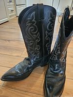 Front view showing black leather boots with decorative white stitching, rounded toes, and pull straps on shafts.