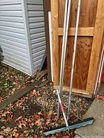 Full view of aluminum handle sections leaning against a wooden wall next to a shed, with the snow shovel blade on the ground