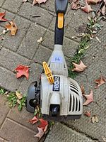 Top view of Ryobi grass trimmer engine showing black and gray casing, yellow throttle lever, and Ryobi branding.