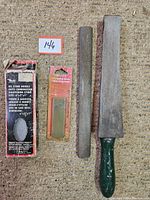 Top view of four sharpening stones/tools laid out on carpet, including an oil stone in box, a sealed small sharpening stone, a round rod stone, and a file with handle.