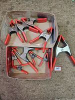 12 spring clamps displayed in two cardboard trays showing variety of sizes; red plastic-coated handles and tips visible