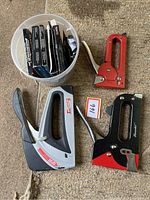 Three manual staple guns and a bucket of staples displayed on carpet, showing condition and size comparison