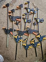 Top-down view of eleven quick-release bar clamps with metal bars and plastic grips in black, blue, orange, and yellow colors, arranged on a carpeted floor.