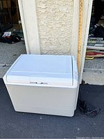 White Coleman electric cooler closed with power cord visible beside it, placed outside near a garage wall with a measuring stick showing height approximate 17.5 inches.