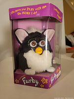 Front view of unopened Generation 1 Skunk Furby in original purple box with clear window showing the black and white plush Furby with pink ears and yellow beak.