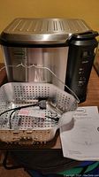 Full view of Butterball electric turkey fryer showing stainless steel body, black control panel, metal frying basket with handle, power cord, and instruction manual.