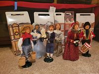 Lot of 7 Byers' Choice Nutcracker caroler figurines standing upright with original illustrated boxes behind them. Figures are dressed in Victorian-style clothing with accessories including birds, cages, drums, and muff. Green bases visible. Some bird accessories show signs of discoloration.