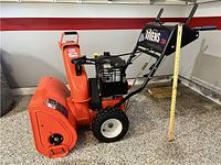Side view of snow blower showing orange housing, 24-inch auger, black engine, wheels, measuring stick showing approx. 41" height.