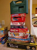 Stack of boxed children's games including UNO Attack, Hedbanz, Twister, Staxis, Mouse Trap, Magic Kingdom and a large Fantasma Magic DVD set box on the floor.