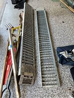 Two metal loading ramps side by side on a garage floor. Each ramp is 76.5 inches long with vented, gripped surfaces, showing dirt and usage marks.