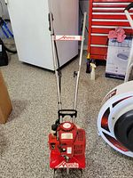 Full front view of red Mantis branded gas-powered tiller showing handle and engine unit on garage floor.
