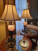Pair of matching table lamps on round wood and glass tables near window with closed curtains. Lamps have faux marble round bases and black metal stands with cream beaded fabric shades.