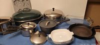 Photo of assorted pots and pans including various sizes of saucepans, cast iron skillet, baking dishes and lids on blue tablecloth