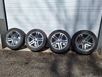 Four 18 inch rims with tires lined up against a garage door, showing the full set.