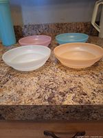 Four pastel colored 6 inch Tupperware cereal bowls (white, pink, blue, peach) displayed on a kitchen counter in a group, showing shape and color.