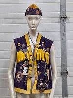 Front view of the purple corduroy vest with yellow trim and fringe, adorned with various enamel pins and badges, and the matching Lions Club hat on a mannequin.