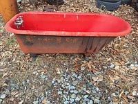 Side view of the red painted cast iron clawfoot bathtub showing original clawfoot feet and rusty exterior at ground level.