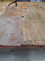 Two worn brown leather aprons laid flat on a wooden workbench showing front and back sides