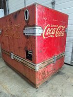 Three angled views of the large mid-century Coca-Cola cooler showing red metal body with yellowed Coca-Cola logos, rusted and worn surfaces, missing side covers and lids, stainless steel liner visible inside, and threaded drain pipe with overflow.