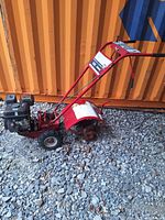 Side view of the Troy-Bilt Junior roto-tiller showing the entire unit including engine, handlebar, and rear tine blades resting on gravel against an orange container.