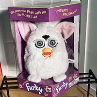 White Furby model 70-800 with green eyes in original purple box with clear front panel. Furby has pink ears, orange beak, and is fluffy.