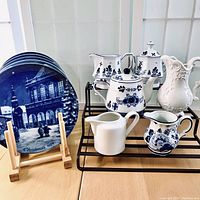 Full lot showing five blue and white German Christmas plates, a white decorative vase pitcher, a floral blue and white creamer and sugar set, a floral teapot, a simple white Fortessa creamer, and a Delft-style mini creamer.