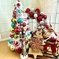 White tinsel Christmas tree decorated with colorful ornaments next to ornate wreath, Santa figurine sitting on chair, and caroler figurines.