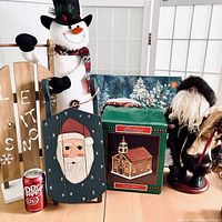 Wide view of all lot items including the Let It Snow wood sign, plush snowman, Old World Santa figure, wooden sled with Santa face, and House of Lloyd Village Church collectible in original box with Dr. Pepper can for scale.