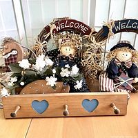 Two Welcome grapevine wreaths with homespun dolls and a wooden peg rack with heart cutouts visible in foreground. Dolls are dressed in star and striped themed outfits, wreaths decorated with straw bows.