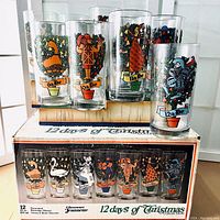 Photo showing 7 of the 12 Jeannette Glass tumblers lined up on original box, showcasing detailed Christmas-themed illustrations for the 6th, 10th, 11th, and 12th days.