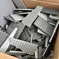 Box containing large assortment of gray LEGO pieces, mainly flat plates in various sizes and shades of gray including light and dark gray.