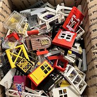 Photo showing mixed LEGO parts including multiple doors, such as red and yellow doors, window frames in white and clear plastic, black and yellow window panels, and some brown doors inside a cardboard box.