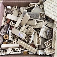 Box of assorted tan LEGO bricks and pieces showing a variety of rectangular and square bricks along with some specialty parts including technic gears and small stickers.