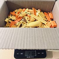 Cardboard box filled with assorted yellow and orange LEGO pieces on digital scale showing 2 lbs weight.