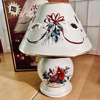 Front view of Lenox Winter Greetings porcelain candle lamp with cardinal image on base and holly and red ribbon decoration on shade, original box behind.