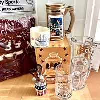 Photo showing a Libbey glass canister in box, large Culver beer stein with colorful artwork, New York Bicentennial ceramic tankard, two Los Angeles Playboy Club glass mugs, Y2K eagle trinket box, golf club covers, old military shirt grippers, and Old Spice shaving mug arranged on a wood table.