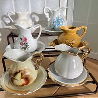 Overview photo showing six sets of mini vintage creamers and saucers of various designs placed on black racks.
