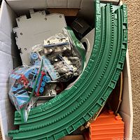 Box with green plastic GeoTrax curved track pieces, roundhouse base, blue and white toy trains in plastic bags