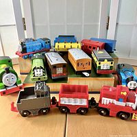 Grouping of 12 Thomas and Friends trains and 3 Fisher Price toy trains displayed on wooden risers and a table, showing various colors and faces of trains.