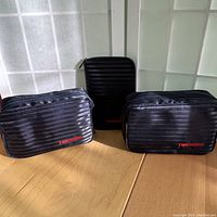 Two black TWA branded toiletry bags with horizontal stripes and zipper closures, plus a small folded expandable black duffel bag.